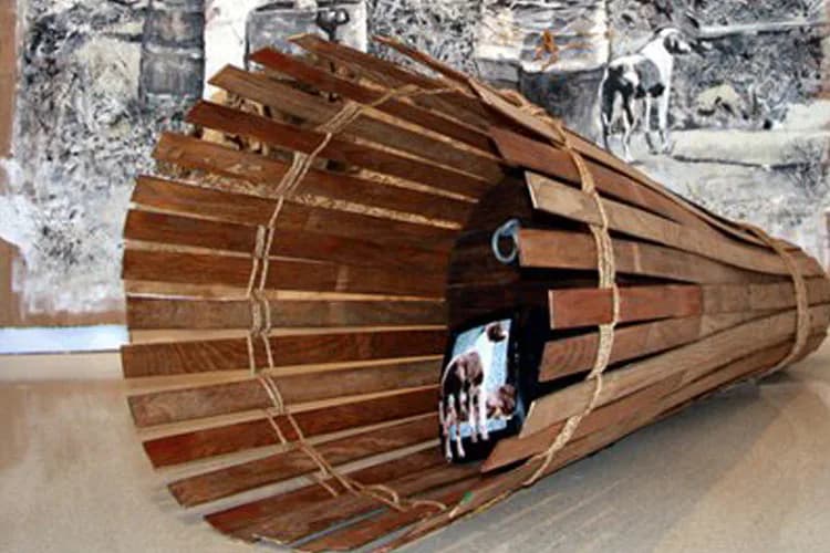 Wooden slat installation sculpture resembling a cone or funnel, bound with rope, displayed indoors with vintage photographs on the wall behind it.