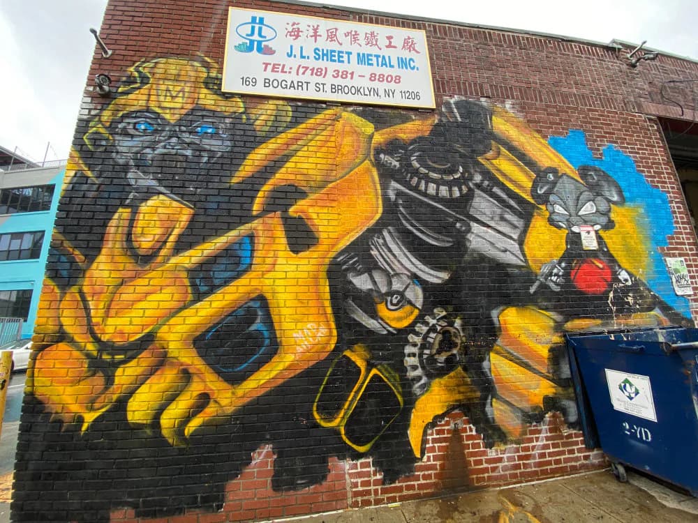 Mad Vaillan mural on brick wall featuring a yellow and black character with geometric shapes and blue accents; spray paint and mixed media.