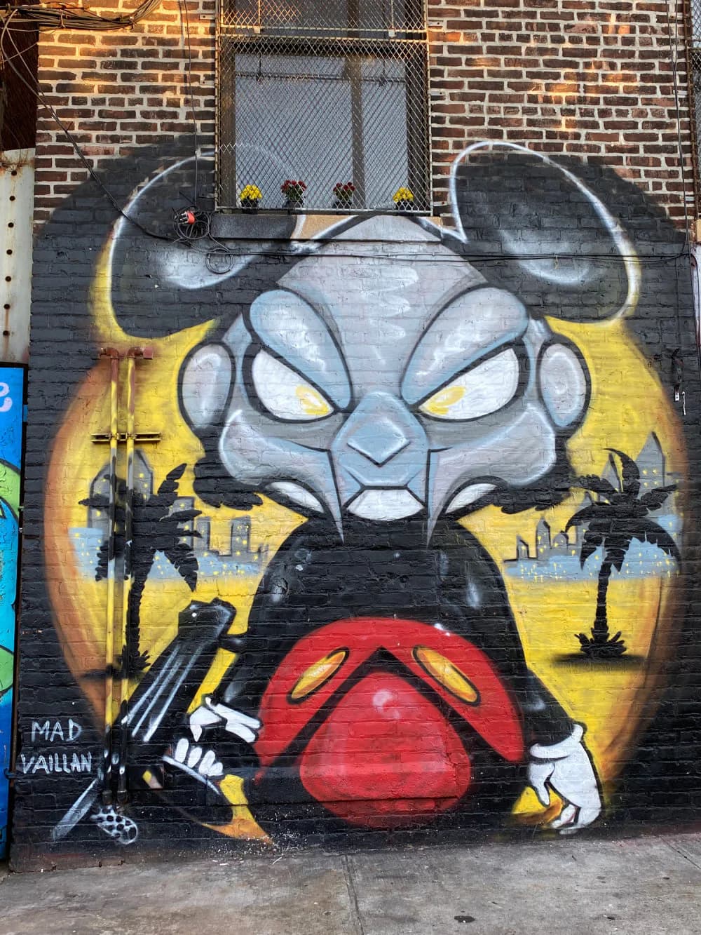 Mad Vaillan mural depicting a gray horned demon head with yellow eyes, red mouth, and white teeth against yellow background with palm trees; spray paint.