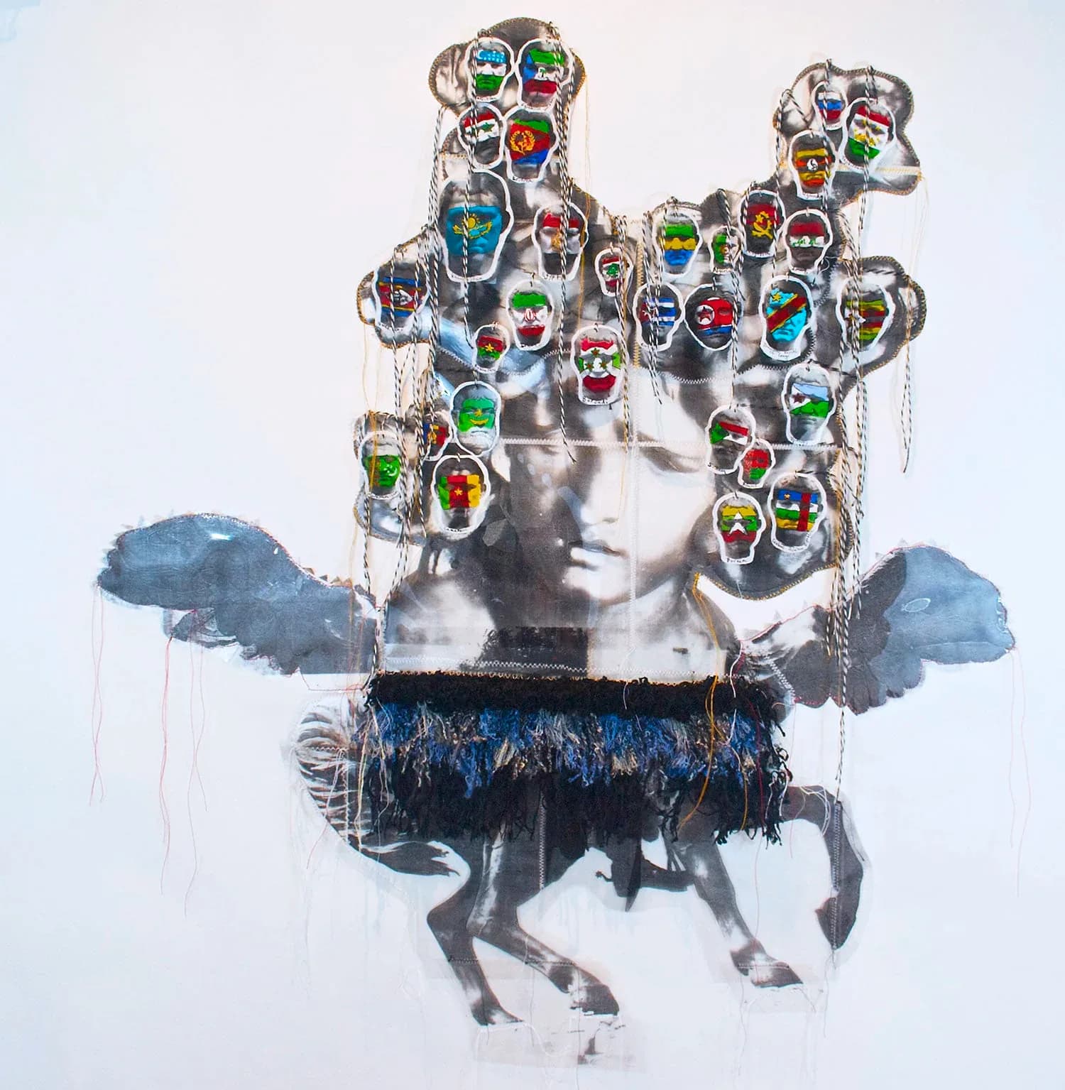 Large mixed-media wall installation of a child figure covered with colorful flag-painted head medallions suspended by chains, with fabric wings and textile fringe