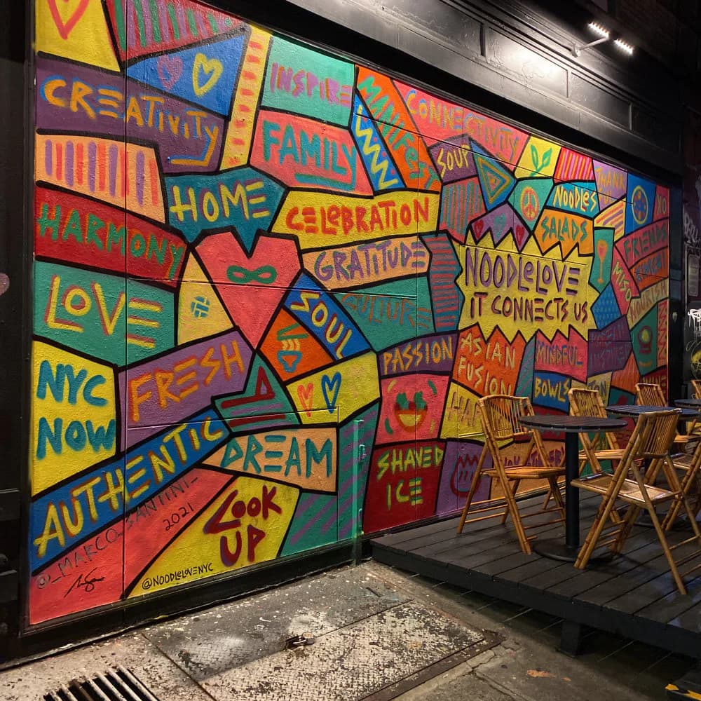 Colorful mural covering a storefront facade with hand-painted words like Creativity, Home, Love, Fresh, and Dream in bold reds, yellows, blues, and greens, with outdoor cafe chairs in front.