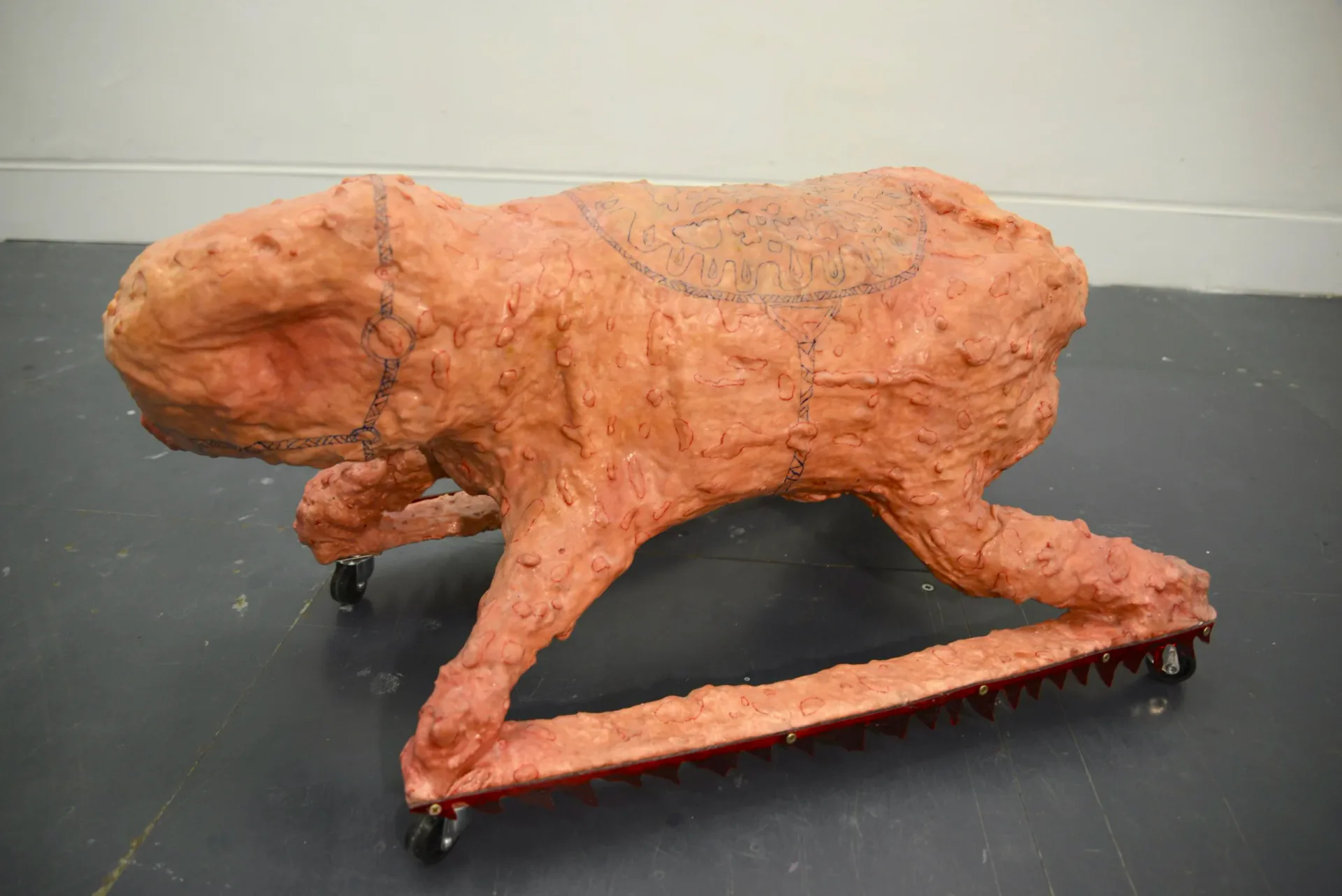 Large flesh-toned animal sculpture with drawn markings, mounted on wheeled metal platform with spikes