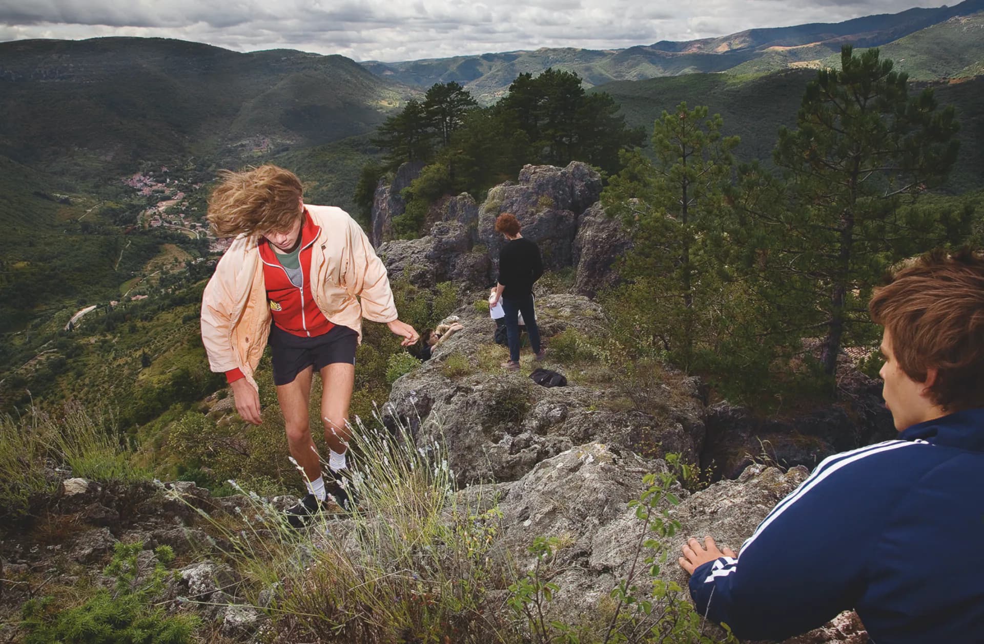 Woman in red and pink jacket running on rocky mountain terrain with green valleys and mountains visible in distant background.