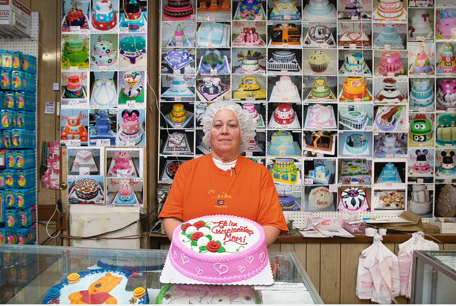 Woman in orange shirt holding a pink frosted cake in front of a wall covered with colorful photographs and cake designs.