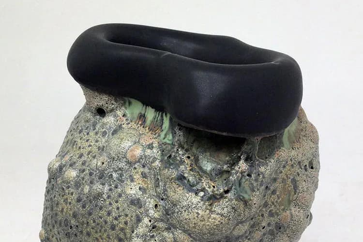 Black rubber or silicone sculptural form resting atop a textured gray stone or concrete base with lichen patches.