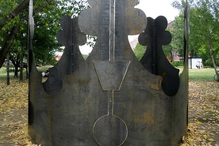 Large weathered metal sculpture with symmetrical organic forms featuring circular openings, displayed outdoors in a park setting.