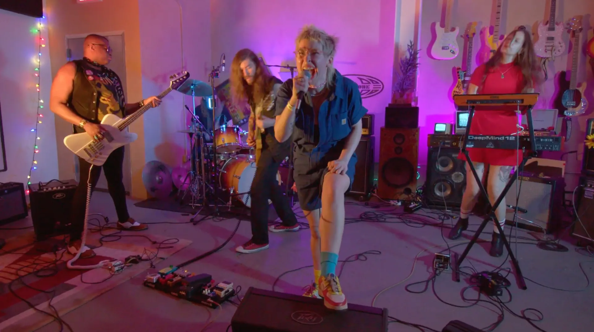 Full band performing in a colorful rehearsal space with guitars, drums, and keyboards