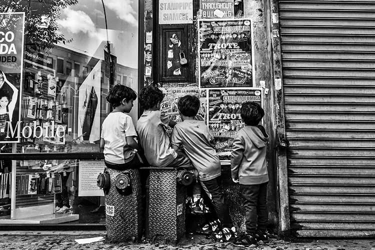 Black and white photograph of four figures from behind, crouched at street level examining a storefront with posters and metal shutters.