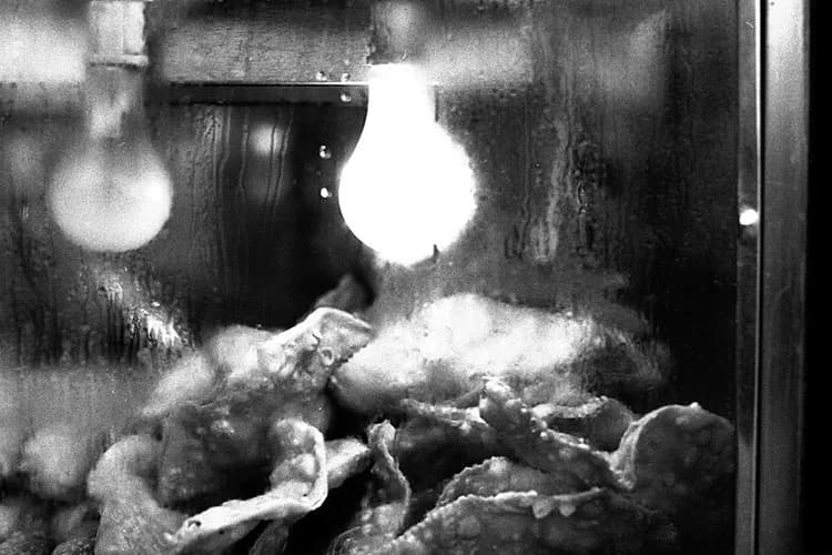 Black and white photograph of a person lying down indoors with bright overhead lights creating stark contrast and shadows; grainy, high-contrast aesthetic.