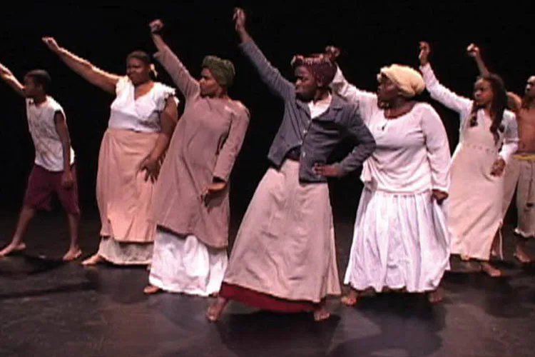 Performance art piece featuring a diverse group of performers on stage wearing pink and white clothing with raised arms in celebratory poses against a dark background.