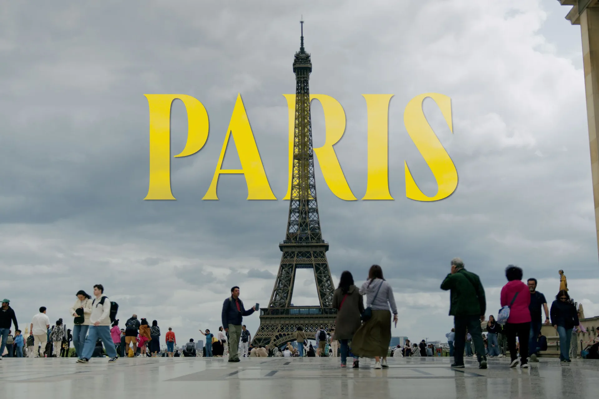 Eiffel Tower with crowds and cloudy sky; large yellow 'PARIS' text overlaid, cinematic title-card style composition.