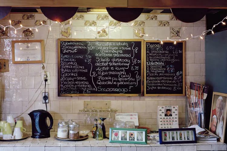 Interior café counter with cream tile backsplash, three chalkboards displaying menu items, kettle, bottles, and kitchen supplies; photograph by Michelle Cheikin.