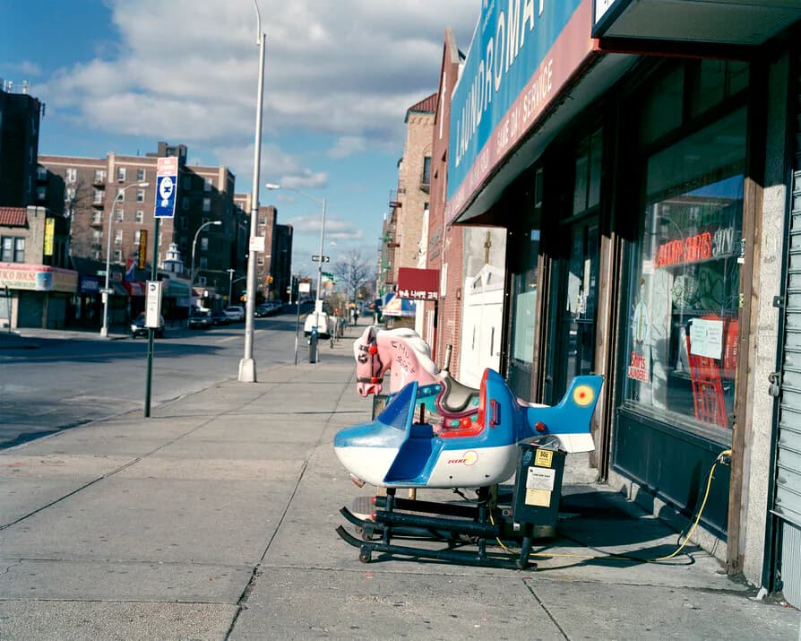 Urban street scene in the Bronx with storefronts, blue sky, and red children's toy vehicle on sidewalk; photograph by Michelle Cheikin.