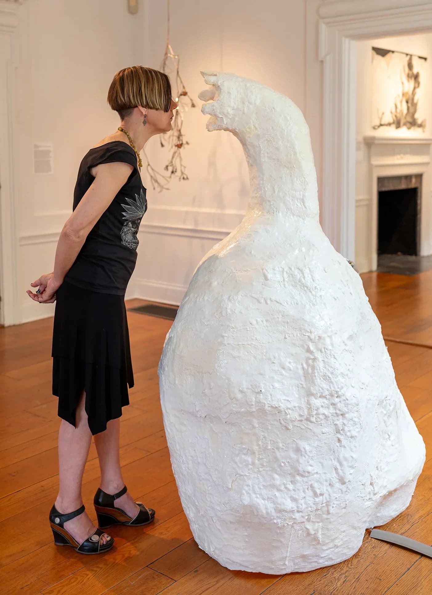 White sculptural figure resembling a goose or swan with an elongated neck, viewed from the side in a gallery space with wood flooring.