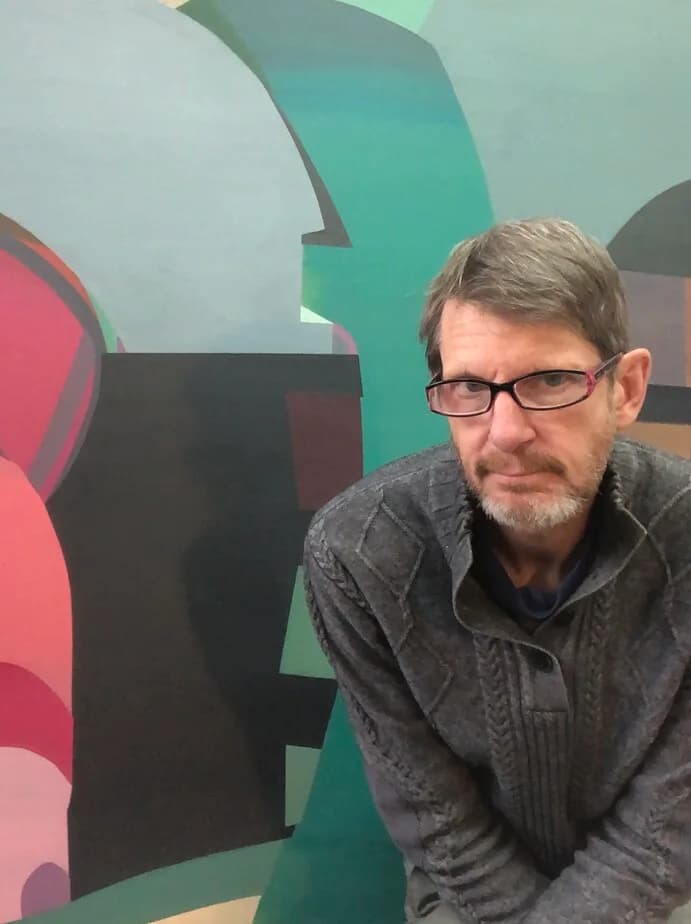 Man wearing glasses and dark jacket photographed indoors, with colorful abstract painted wall in red, teal, and green behind him.
