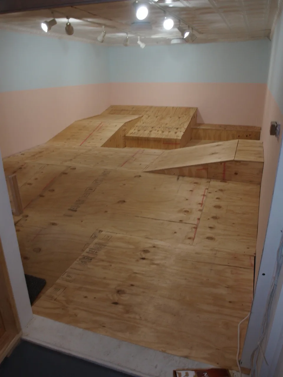 Plywood sculptural floor installation with ramps and platforms in gallery with pink and blue painted walls