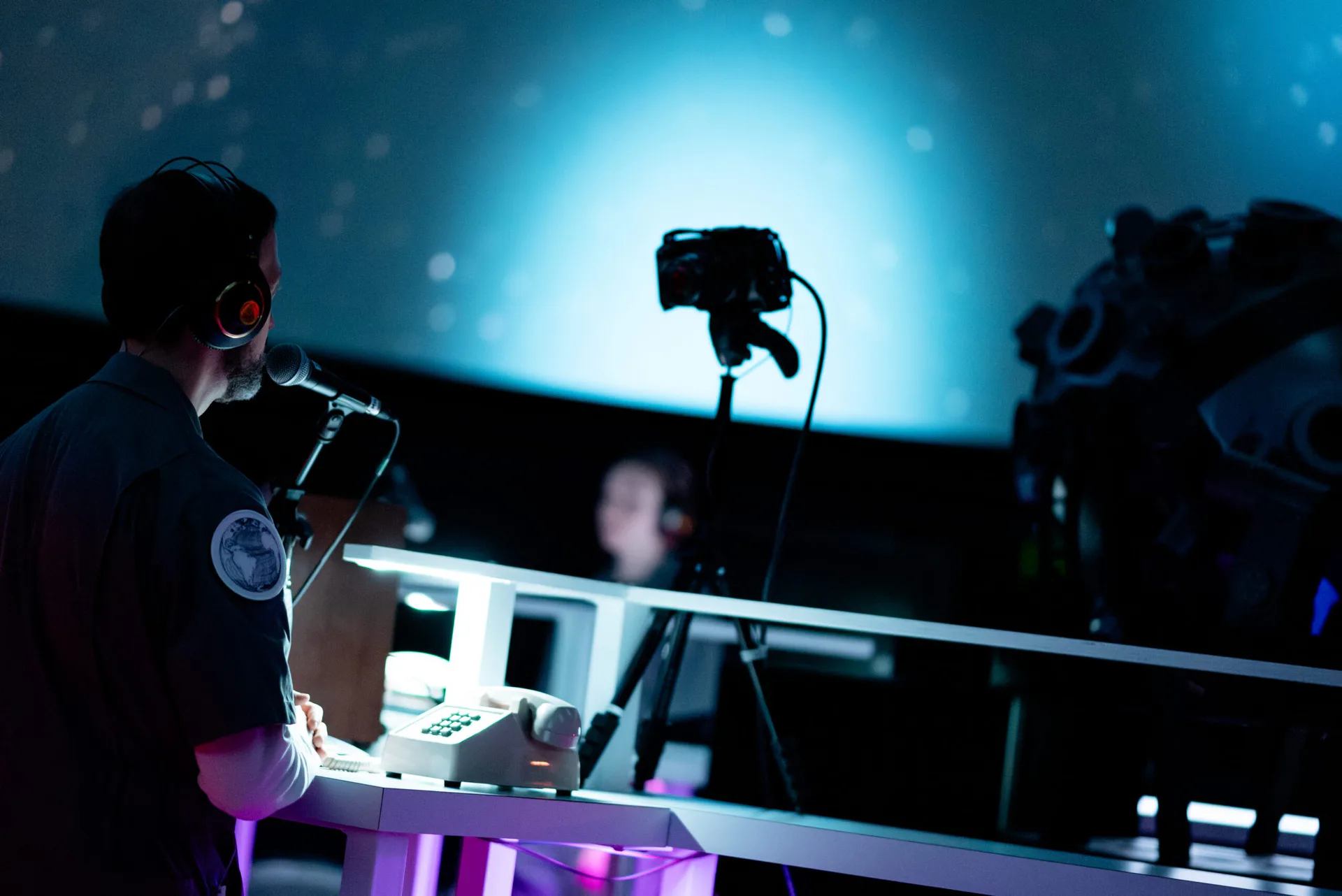 Performer kneeling and singing beneath a NASA spacesuit projected on ceiling in darkened theater