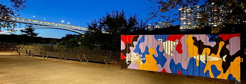 Outdoor urban setting with colorful abstract animal mural on shipping container, green space, and city bridge and skyline in background.
