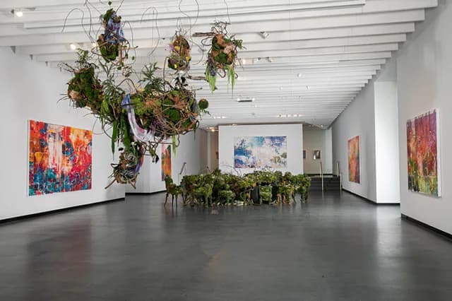Modern gallery interior with white walls and ceiling, featuring suspended hanging plant installation and colorful abstract paintings.