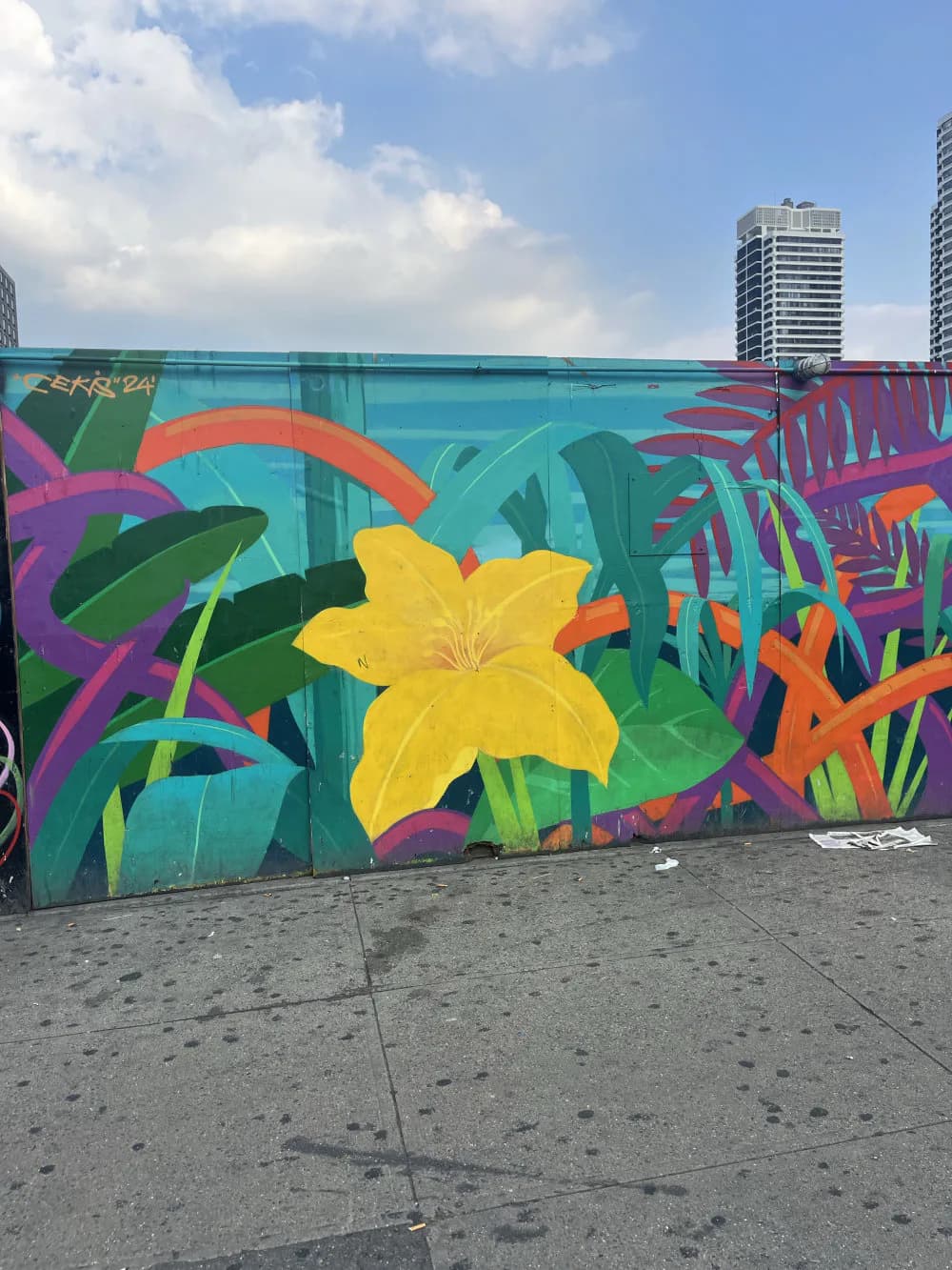 Vibrant mural featuring tropical flowers and foliage in yellow, orange, pink, and teal on turquoise background, painted on urban wall.