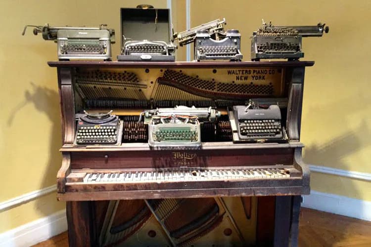 Mixed-media installation featuring vintage typewriters arranged on and around a wooden piano, with sheet music visible, creating a sculptural assemblage combining musical and writing instruments.
