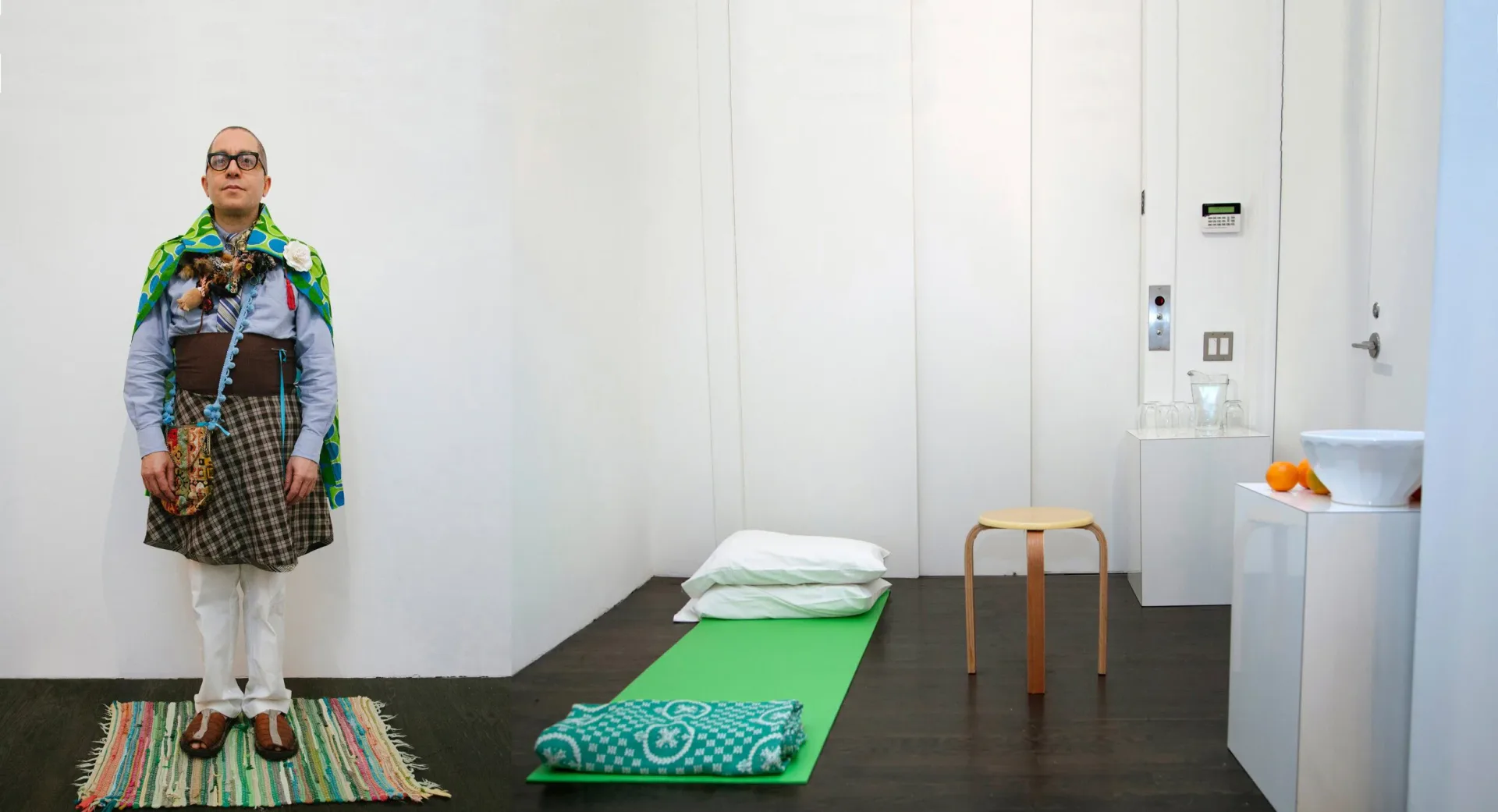 Mixed-media installation with a standing figure in colorful patterned clothing, green bedding on black platform, and white room interior.