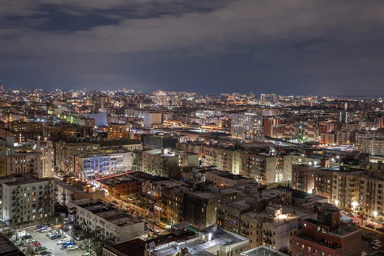 Nighttime cityscape photograph of the Bronx with illuminated buildings, streets, and residential structures under a dramatic cloudy sky.