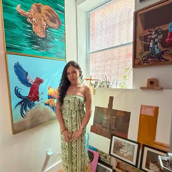 Artist Pamela Baloyo stands in bright studio with colorful framed paintings on walls including sea turtle on turquoise, rooster on blue, and other artwork.