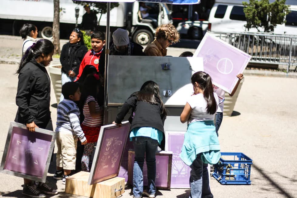 Children and adults gather around a printmaking screen station outdoors, holding purple screen-printed posters near a van, related to Patrick Rowe's community print project