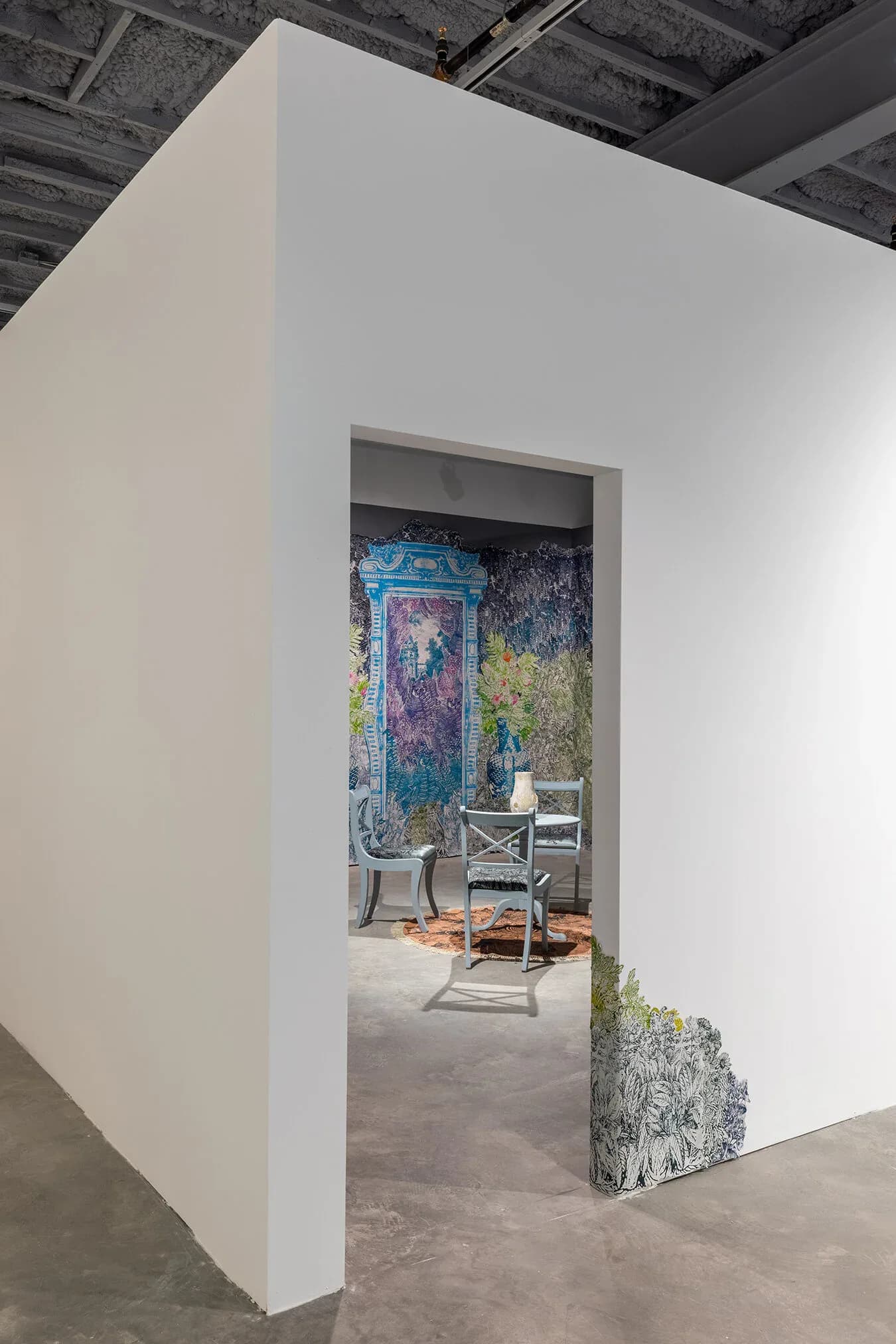 Installation view through white doorway framing painted backdrop with floral and textural patterns, chair and decorative elements visible inside.