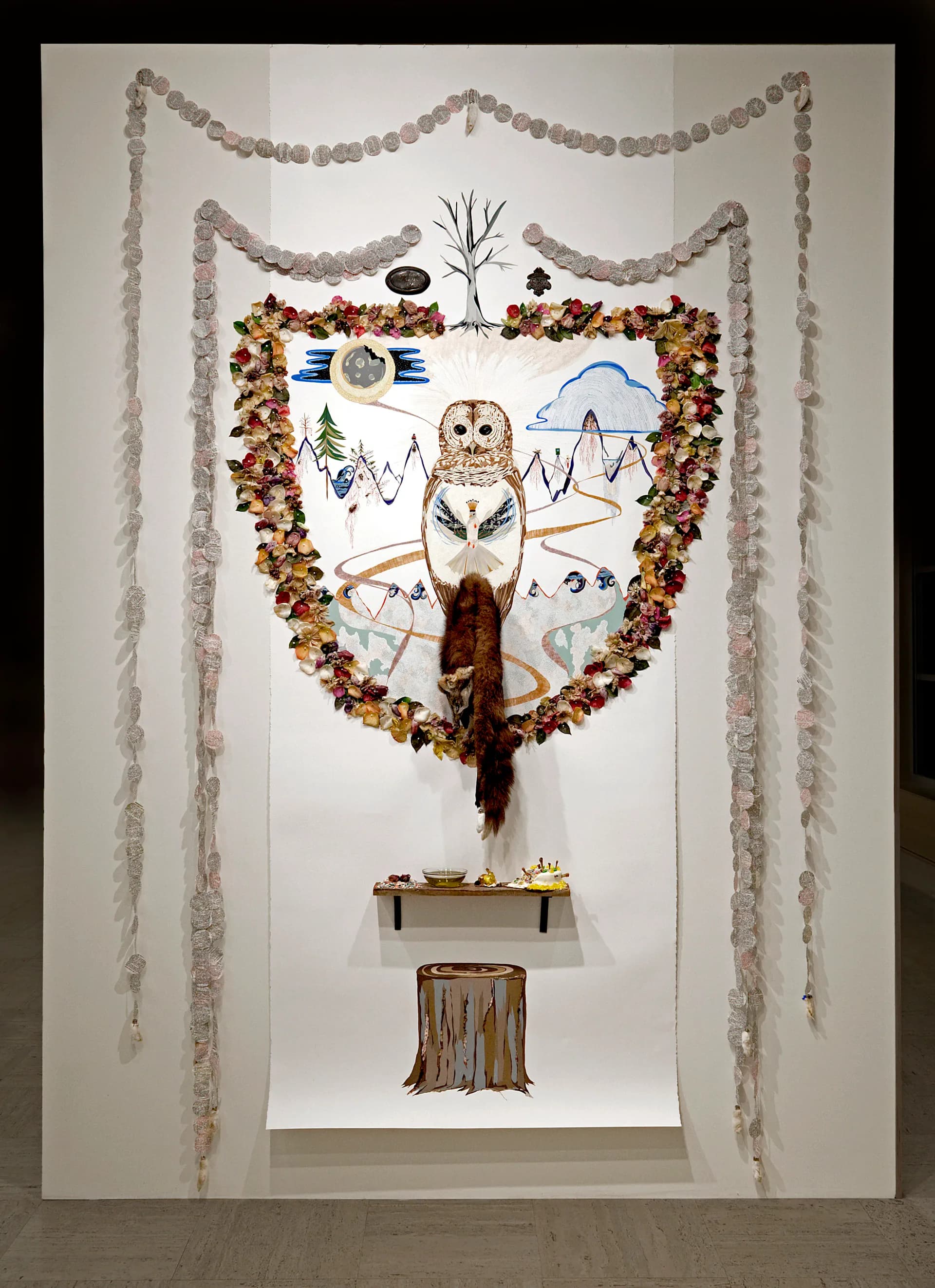 Large mixed-media installation featuring a barred owl drawing surrounded by dried flowers, paper discs, and found objects on a gallery wall