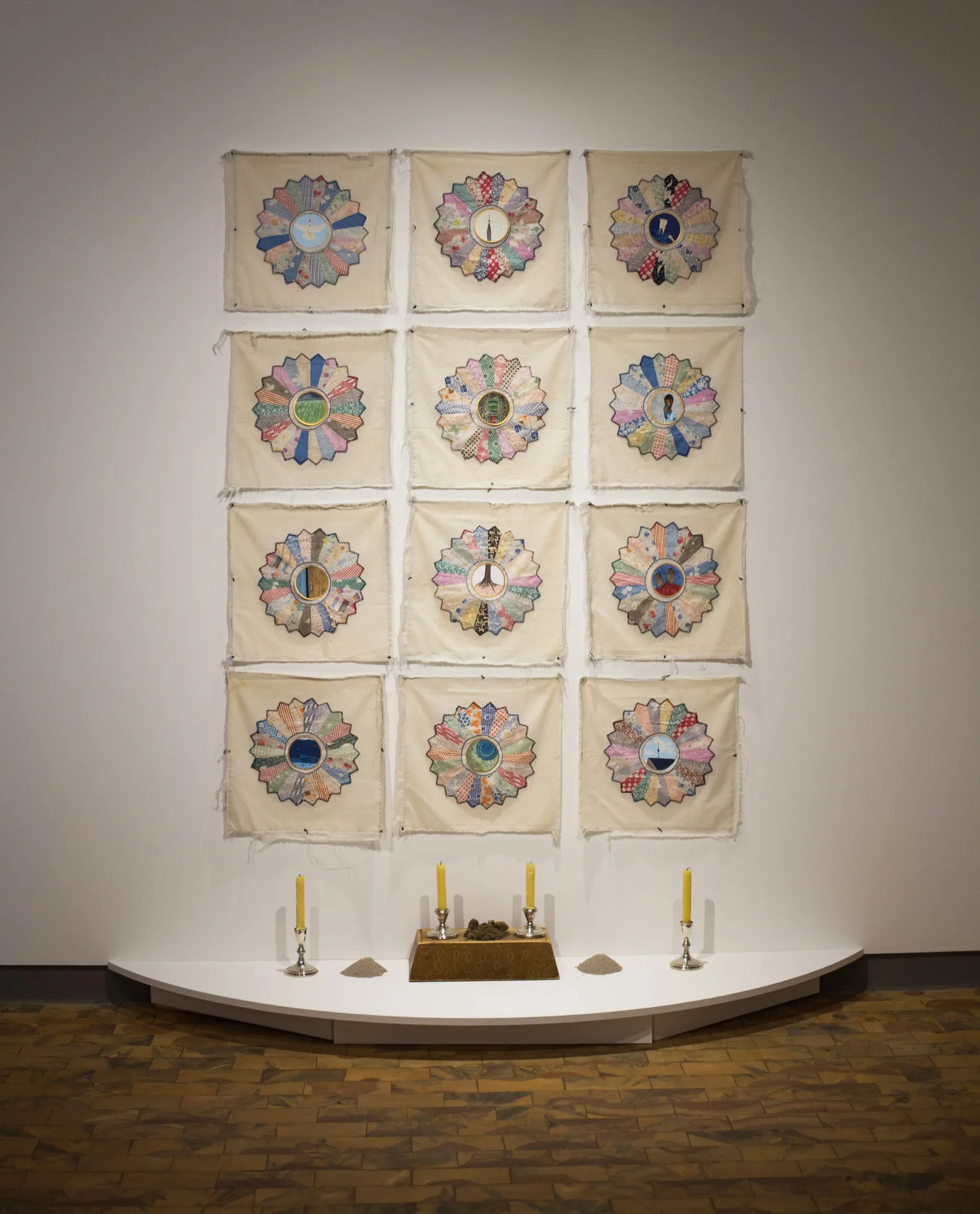 Grid of twelve quilt-based artworks with painted circular medallions, displayed on a gallery wall above a candle altar