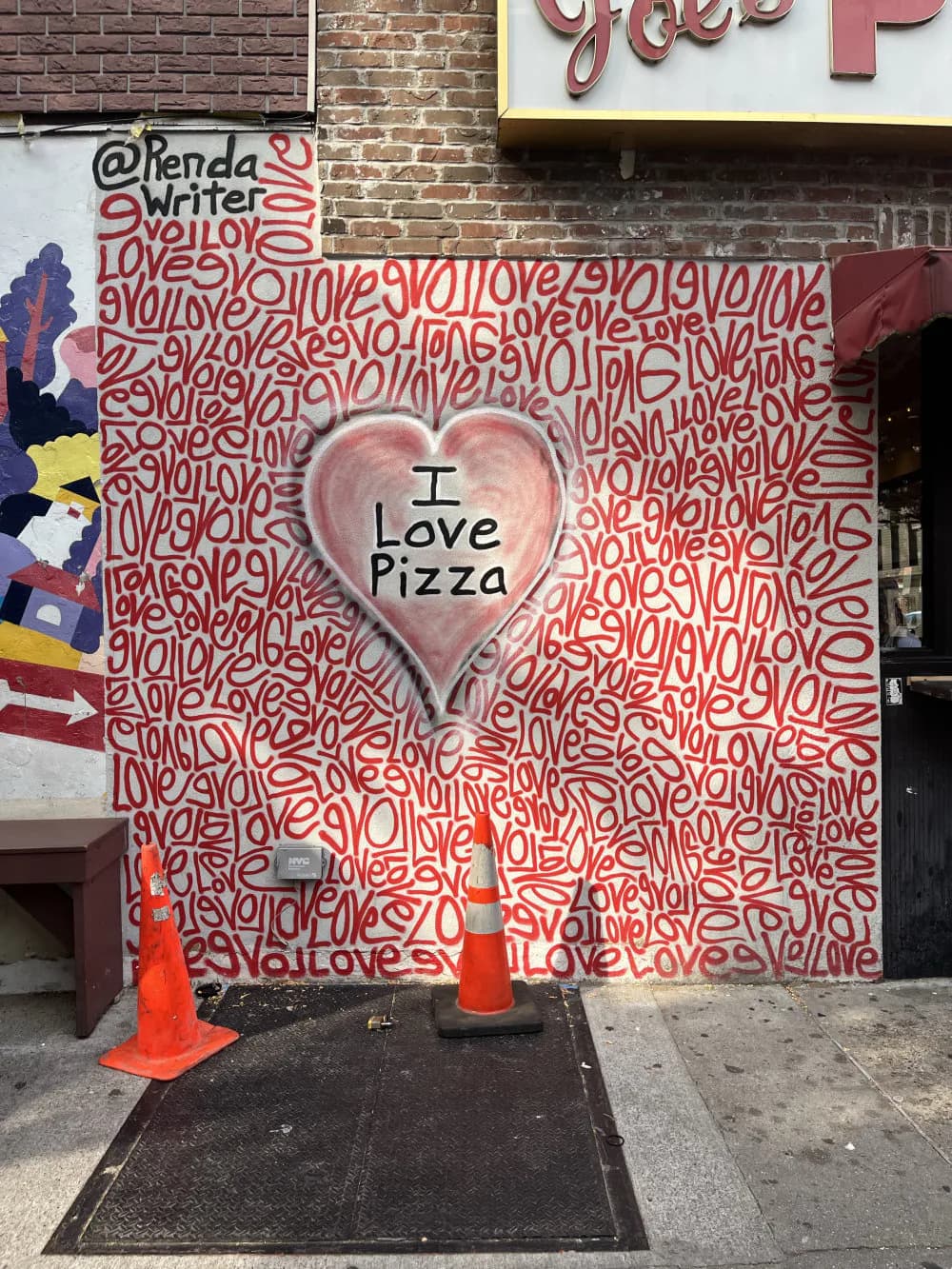 Mural with red and white repeating text pattern background and large pink heart containing "I Love Pizza" text, flanked by orange traffic cones.