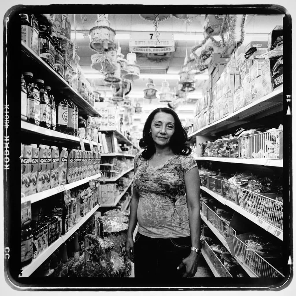Black and white photograph of a pregnant woman standing in a supermarket aisle between shelves of products, looking directly at camera.