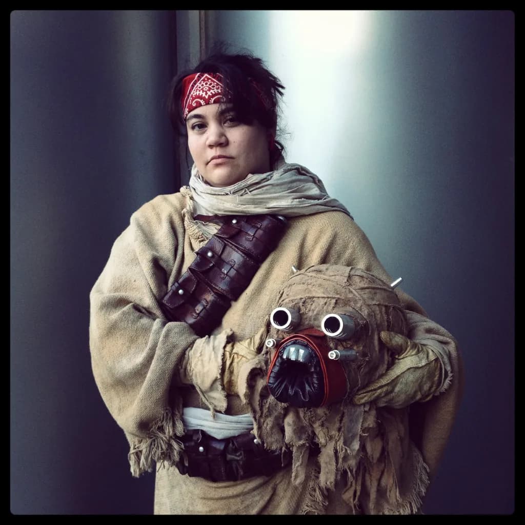 Portrait photograph of a person wearing a beige jacket and headwrap, holding a sculptural creature prop with mechanical details against a gradient background.