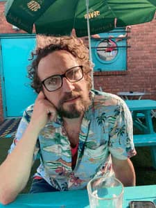 Color photograph of a man with curly hair and glasses wearing a tropical-print shirt, seated outdoors beneath a green umbrella.