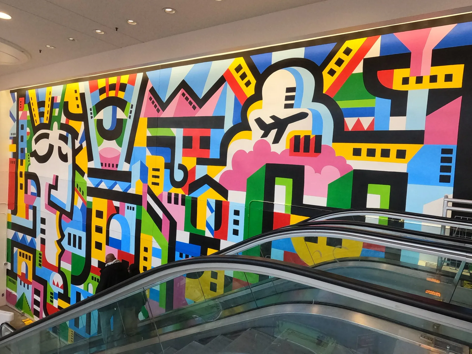 Vibrant abstract mural installation with bold geometric shapes, primary colors, and playful figures on an interior gallery wall.