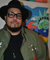 Portrait of Rob Ayala wearing glasses and an olive green jacket over black, posed in front of a colorful abstract painting.