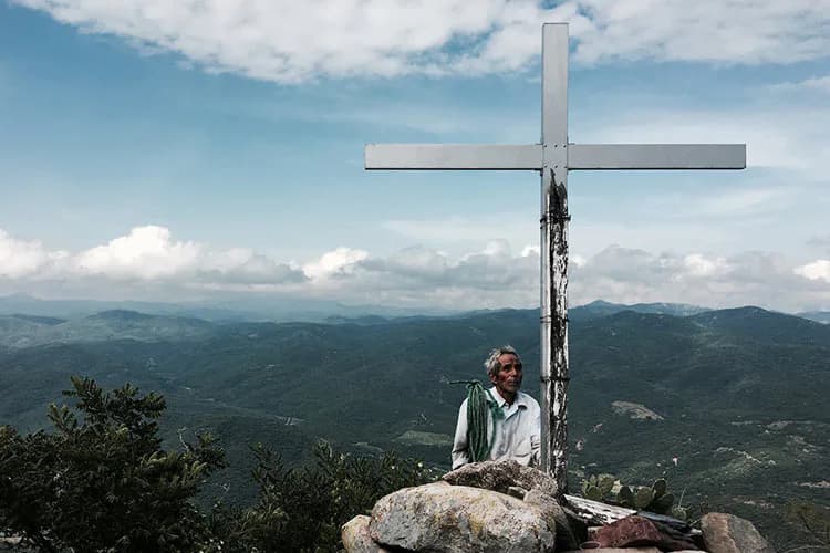 Photograph of a person standing beside a large white cross on a rocky mountain overlook, with forested valleys and distant mountains under blue sky with white clouds.