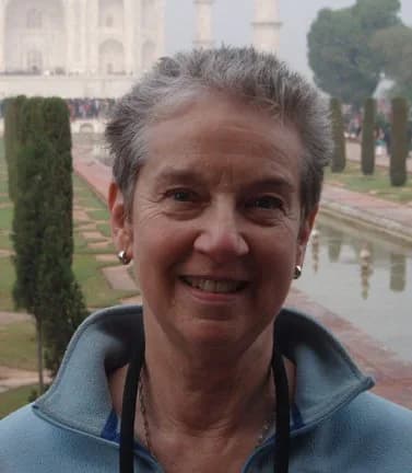 Smiling woman with gray hair wearing a blue jacket, photographed outdoors with a white building and cypress trees in the background.