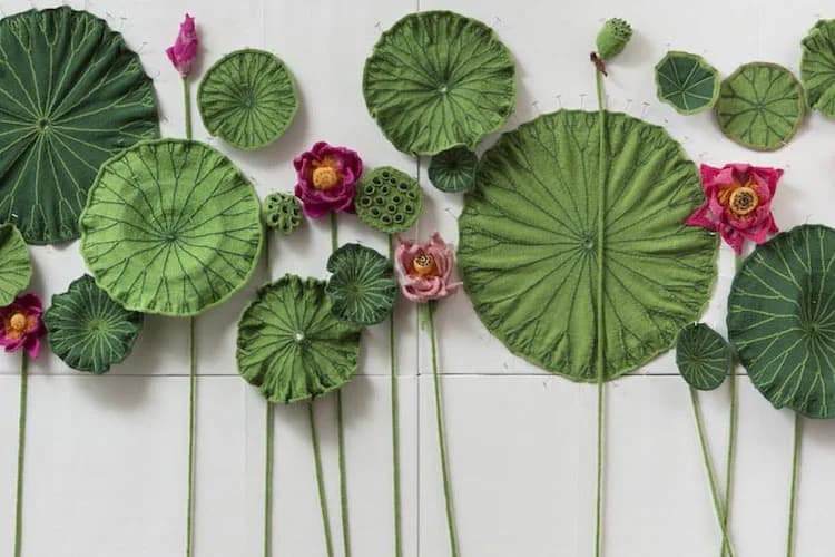 Installation artwork featuring green lily pad leaves of varying sizes arranged against white wall with pink and magenta lotus flowers interspersed among green stems.