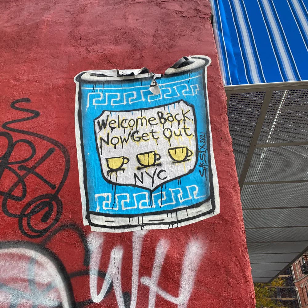 Street art mural on red wall featuring blue can-shaped design with Greek pattern border, "Welcome Back You Get Out" text, and yellow coffee cup illustrations.
