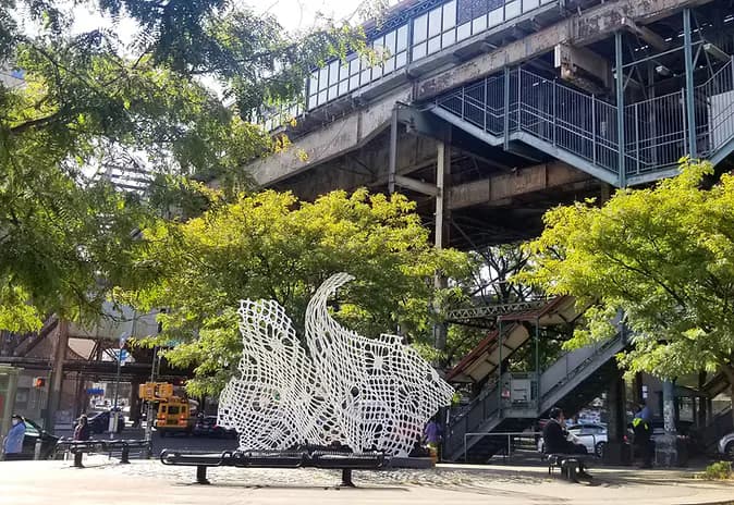 Outdoor public plaza beneath a concrete overpass with green trees, featuring a large white geometric sculptural installation by Samantha Holmes on a dark platform.