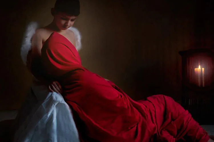 Woman with white wings wearing red fabric and hat, posed dramatically against dark background with candlelight.