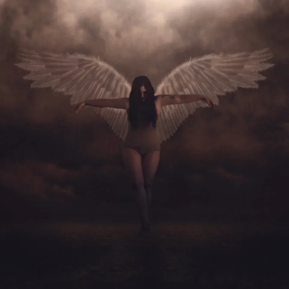 Woman with long dark hair and large white wings spread wide, standing in misty atmosphere against clouded sky.