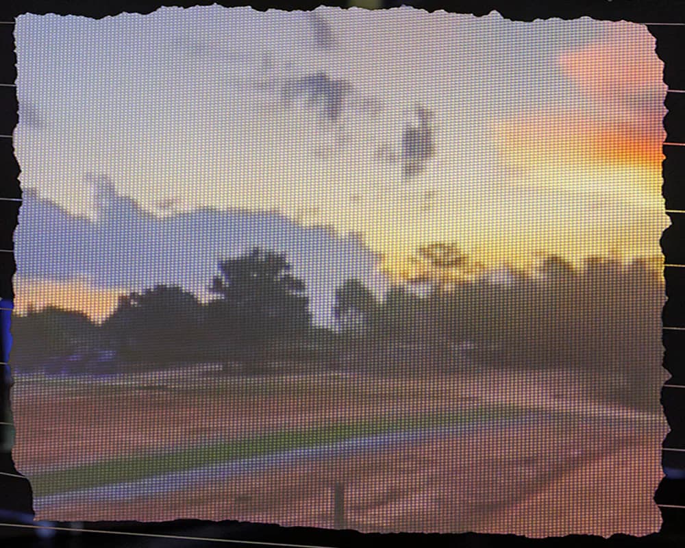 Pixelated or digitized landscape showing a sunset over fields with trees, rendered in soft pastels and warm orange tones with visible compression artifacts.