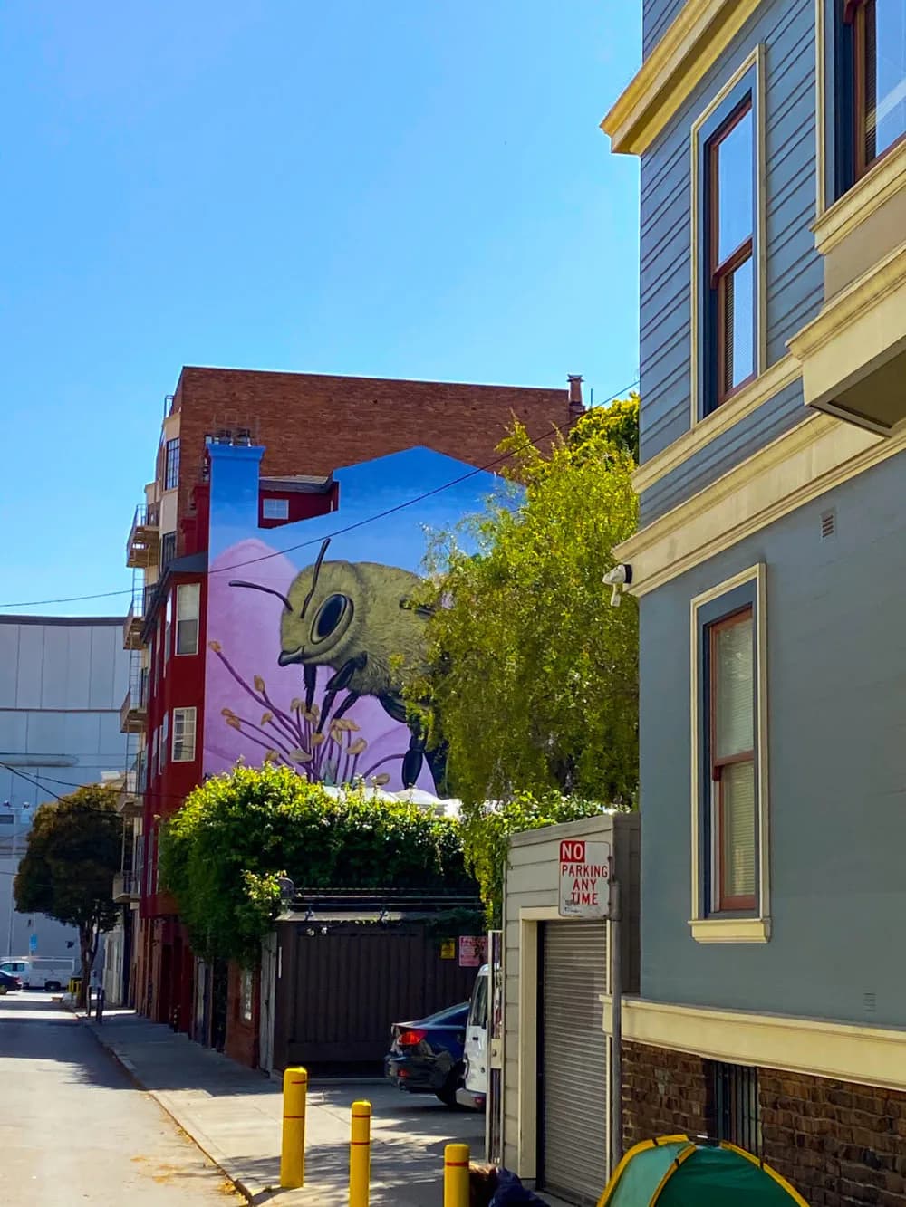 Photograph of urban street with brick buildings, a colorful mural featuring purple and blue tones on a building wall, green foliage, and yellow bollards.