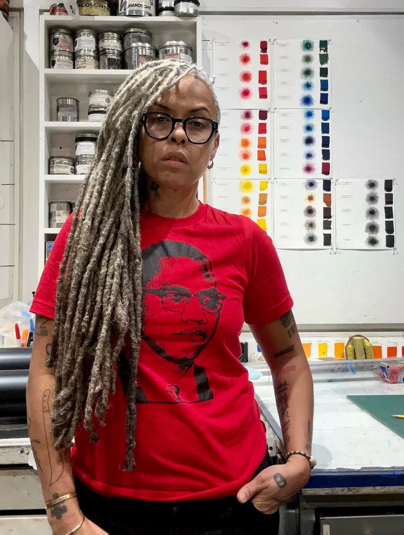 Person with long gray dreadlocks wearing red graphic t-shirt and glasses in an art studio with color swatches displayed behind them.