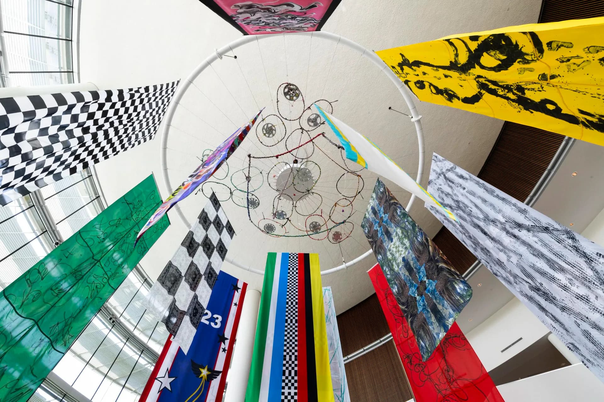 Large-scale installation with painted banners and bicycle wheels suspended from a circular frame in an atrium