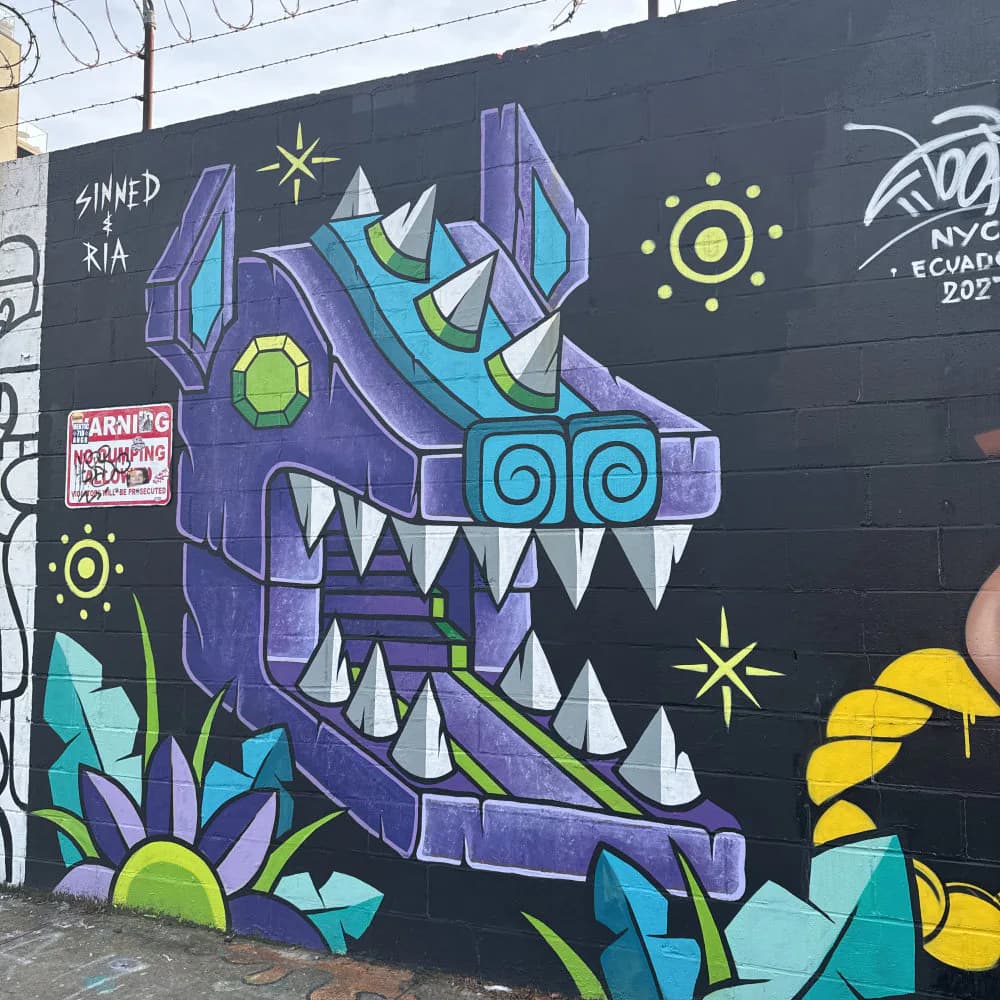 Street mural by Sinned featuring a purple geometric monster head with sharp teeth, cyan accents, and yellow orbital elements on black background.